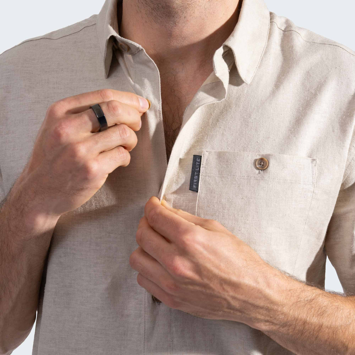 Man adjusting the collar of a beige shirt with a First Lite label on a light background