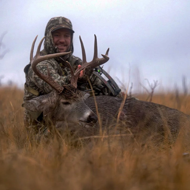 Smiling hunter in camouflage, with bow, next to a large 8-point buck in tall grass.