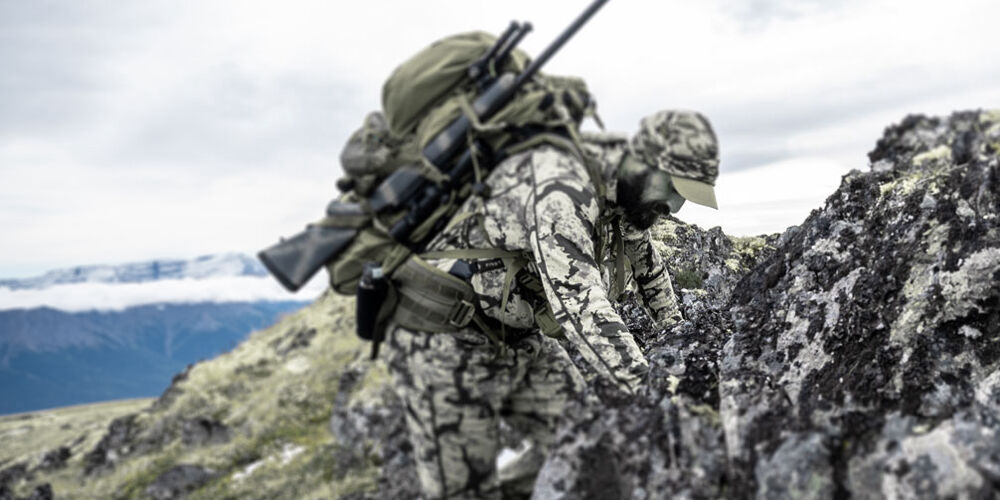 Person in grey camouflage outfit and olive backpack with rifle ascends rocky terrain.