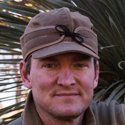 Tan cap with ear flaps and black string bow on front.