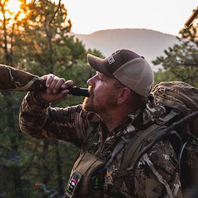 Hunter blowing a black game call with a camouflage hose outdoors.