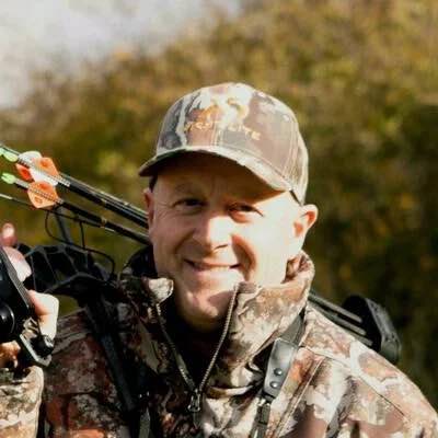 Smiling man in camouflage hunting gear carrying a bow with orange arrows.