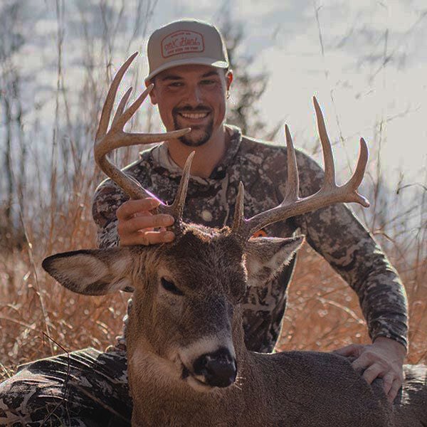Smiling hunter in tan cap and camo shirt with large antlered buck.