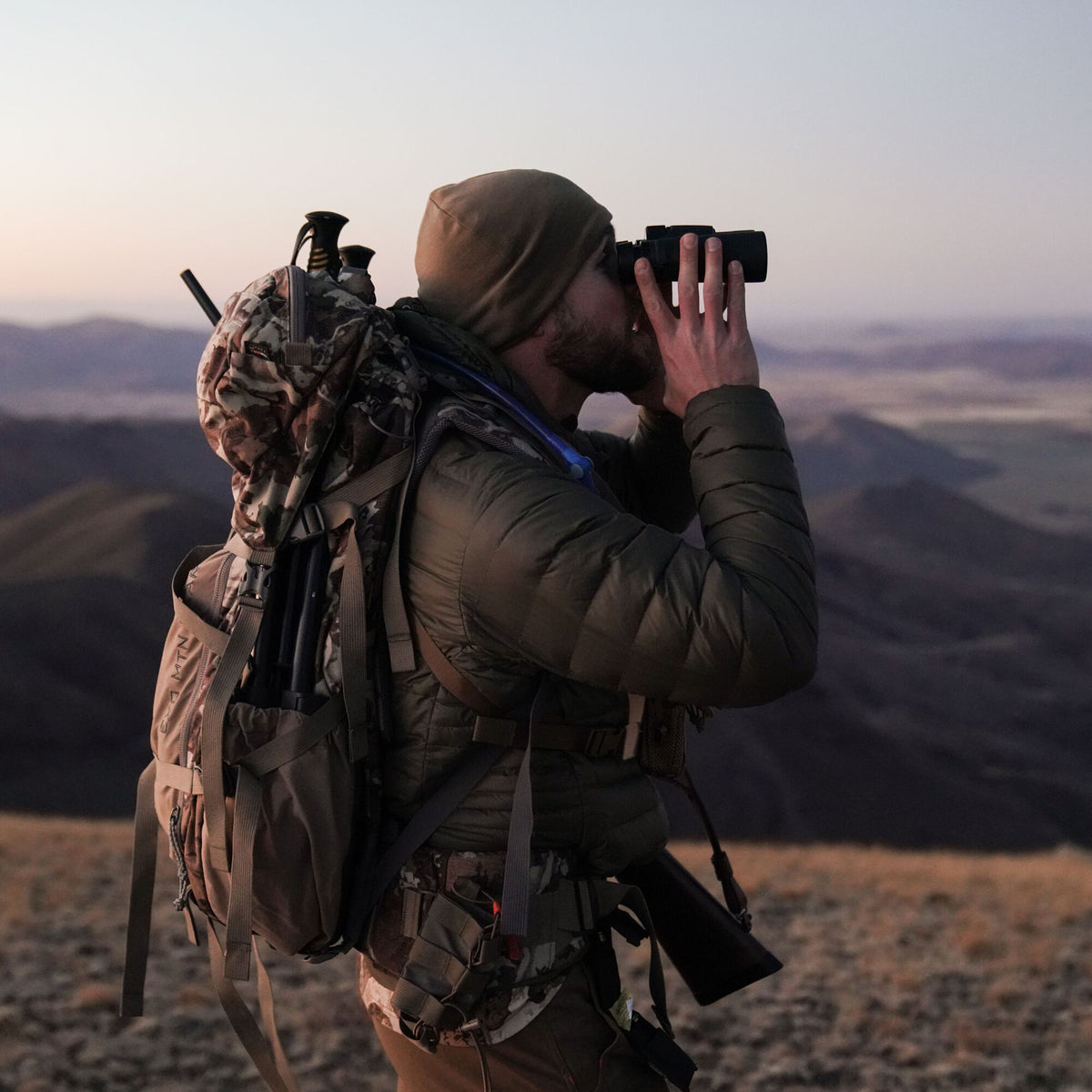A man in an olive green puffer jacket and camouflage EXO MTN backpack with trekking poles and a rifle scans mountain scenery with black binoculars.