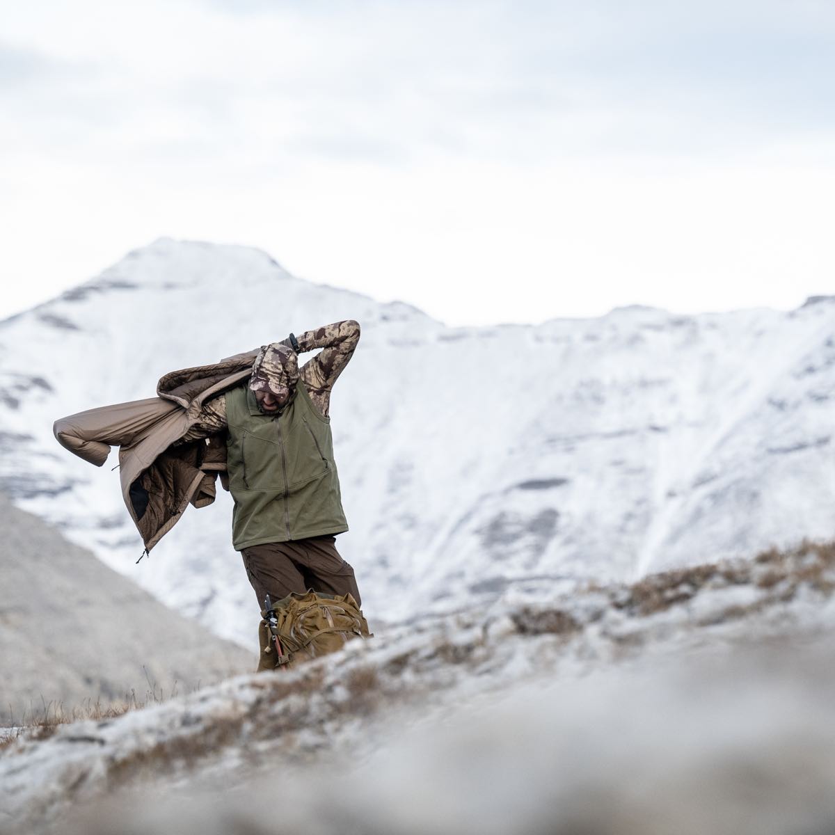Man adjusting tan insulated jacket over olive vest in snowy mountains.