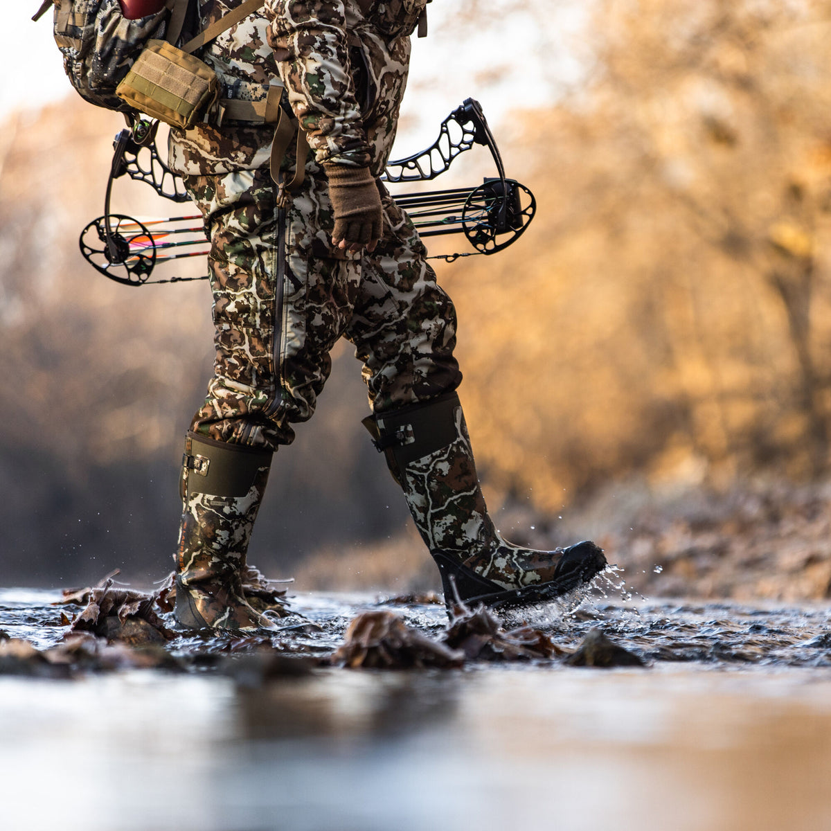 Tall camouflage rubber hunting boots with a dark buckle strap, splashing water as a hunter wades.