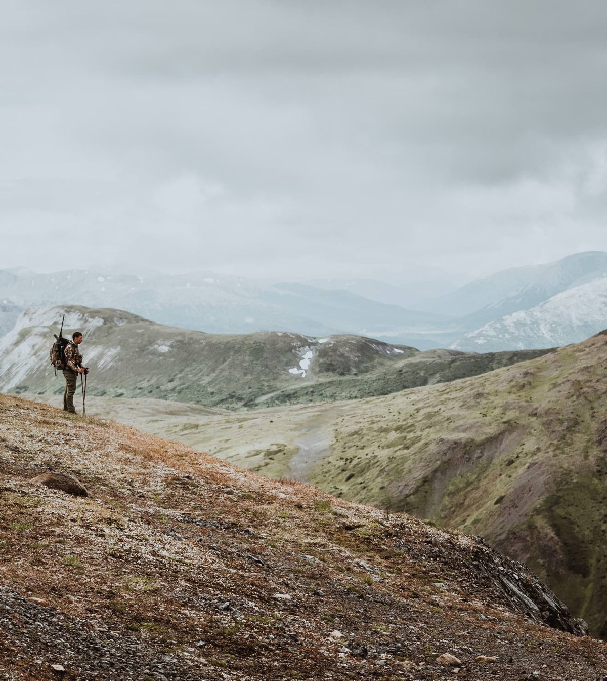 Man in camouflage with rifle-mounted backpack and trekking poles on mountain.