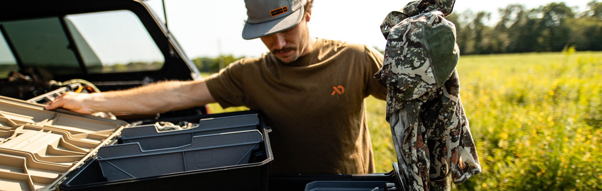 Man loads tan and dark grey modular storage system into truck bed, holding camo gear.