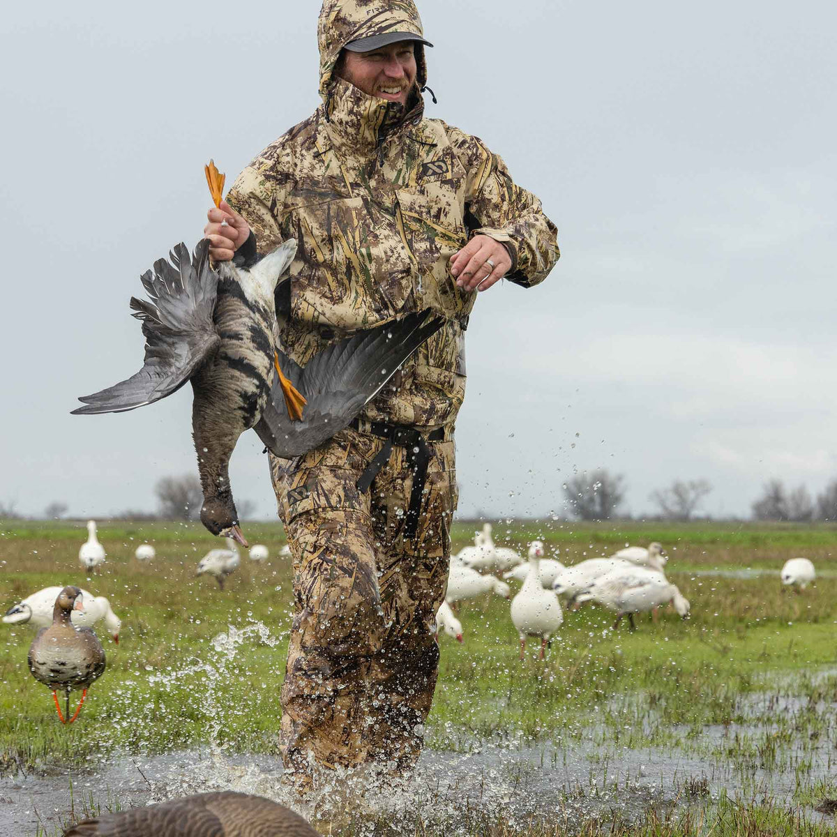 A man in a hooded, earth-toned camouflage hunting suit with a visible chest logo splashes through shallow water while holding a goose.