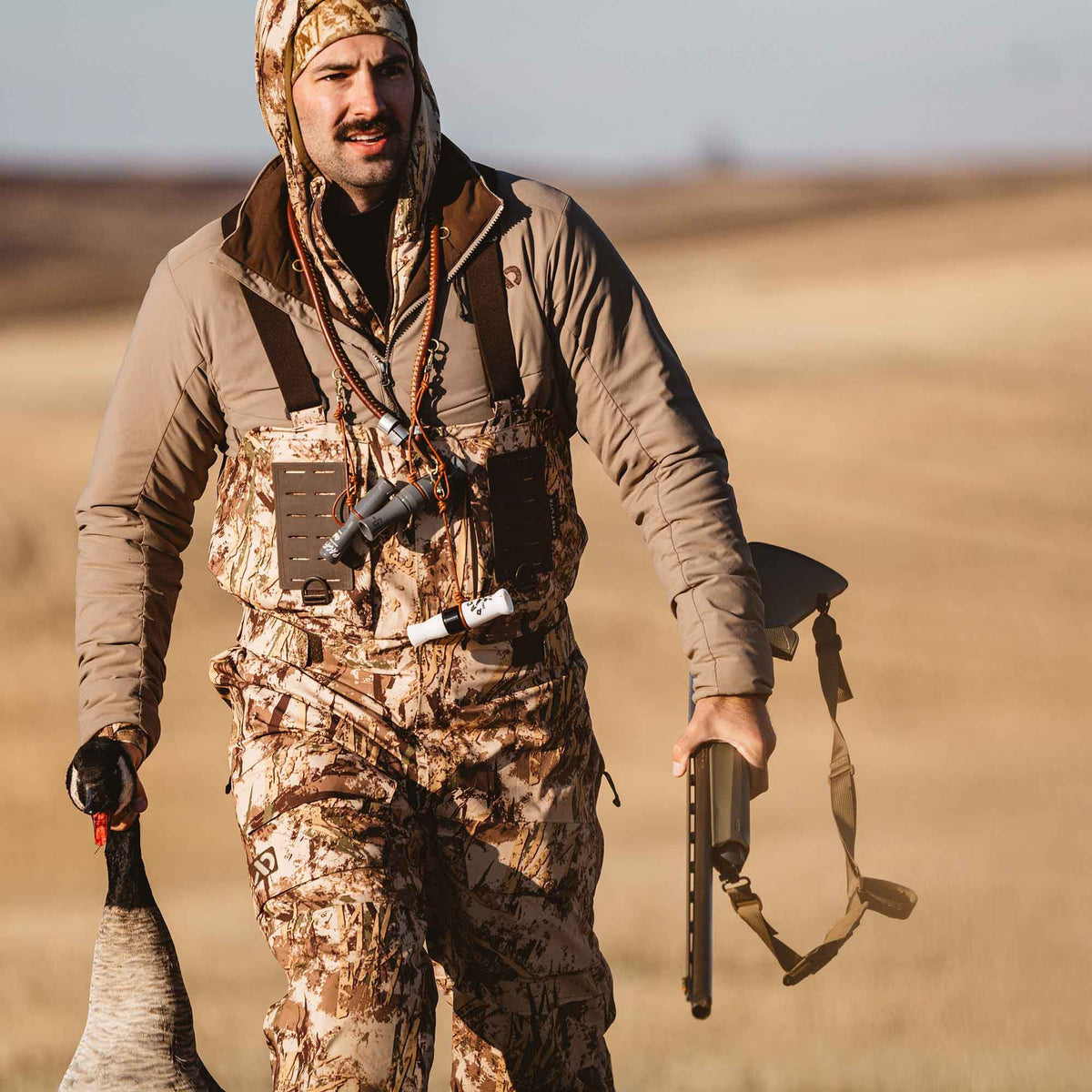 A hunter wears a tan jacket and brown-tan camouflage waders with a hood and suspenders, carrying a shotgun and a goose.