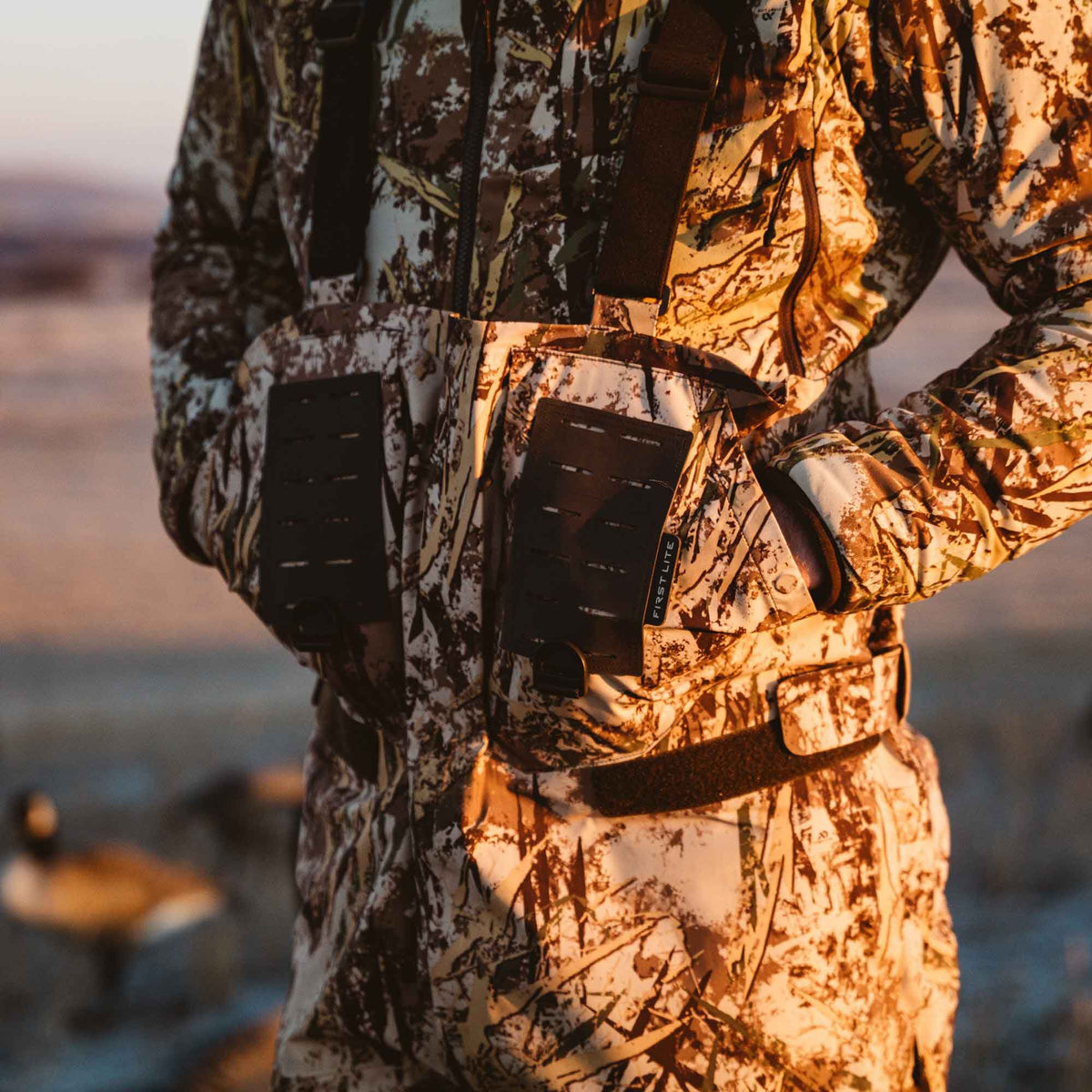 Person wearing brown, white, and green camouflage hunting bibs featuring black modular chest panels with a First Lite logo.