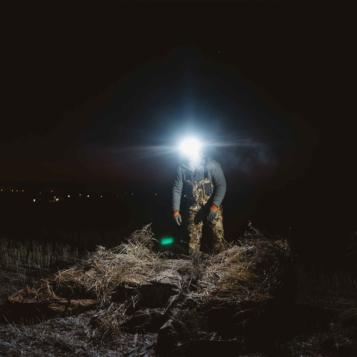 A person in camouflage waders and orange gloves wears a bright white headlamp, standing in a dark, grassy field.