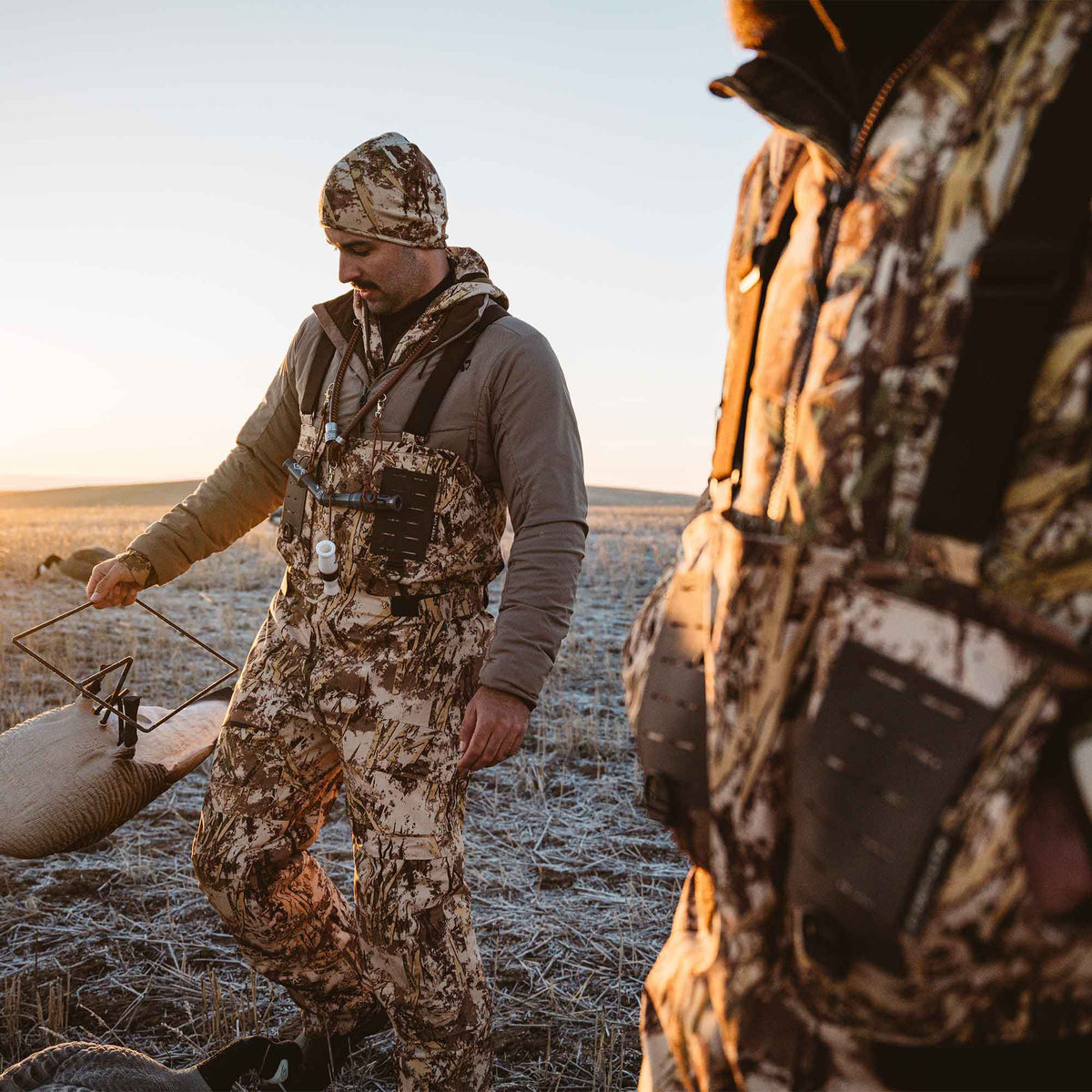A hunter wears tan and brown wetland camouflage hunting bibs with attached calls, a matching beanie, and a solid tan jacket.