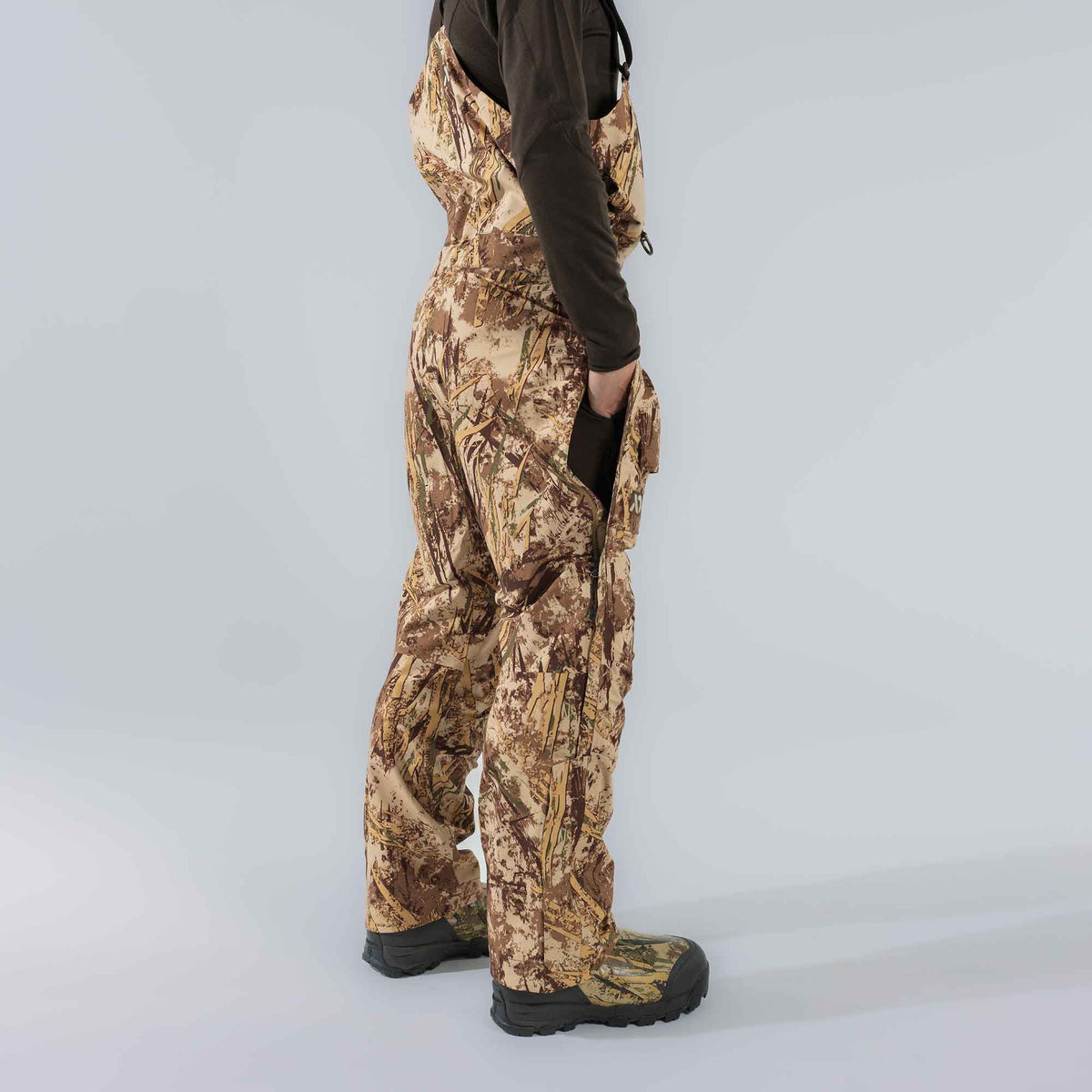 Person wearing tan and brown marsh camouflage waders with suspenders, a side zipper partially open at the hip, and integrated boots.