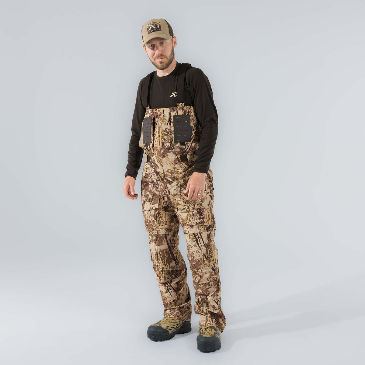 A man models tan and brown camouflage hunting waders with integrated black boots and chest utility panels.