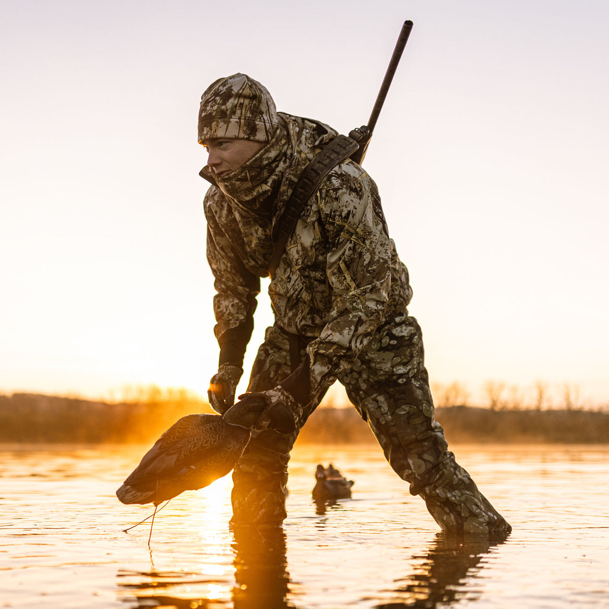 Hunter in beige and brown camouflage hunting suit, beanie, and neck gaiter, holding a duck decoy and carrying a shotgun, wading in sunrise-lit water.