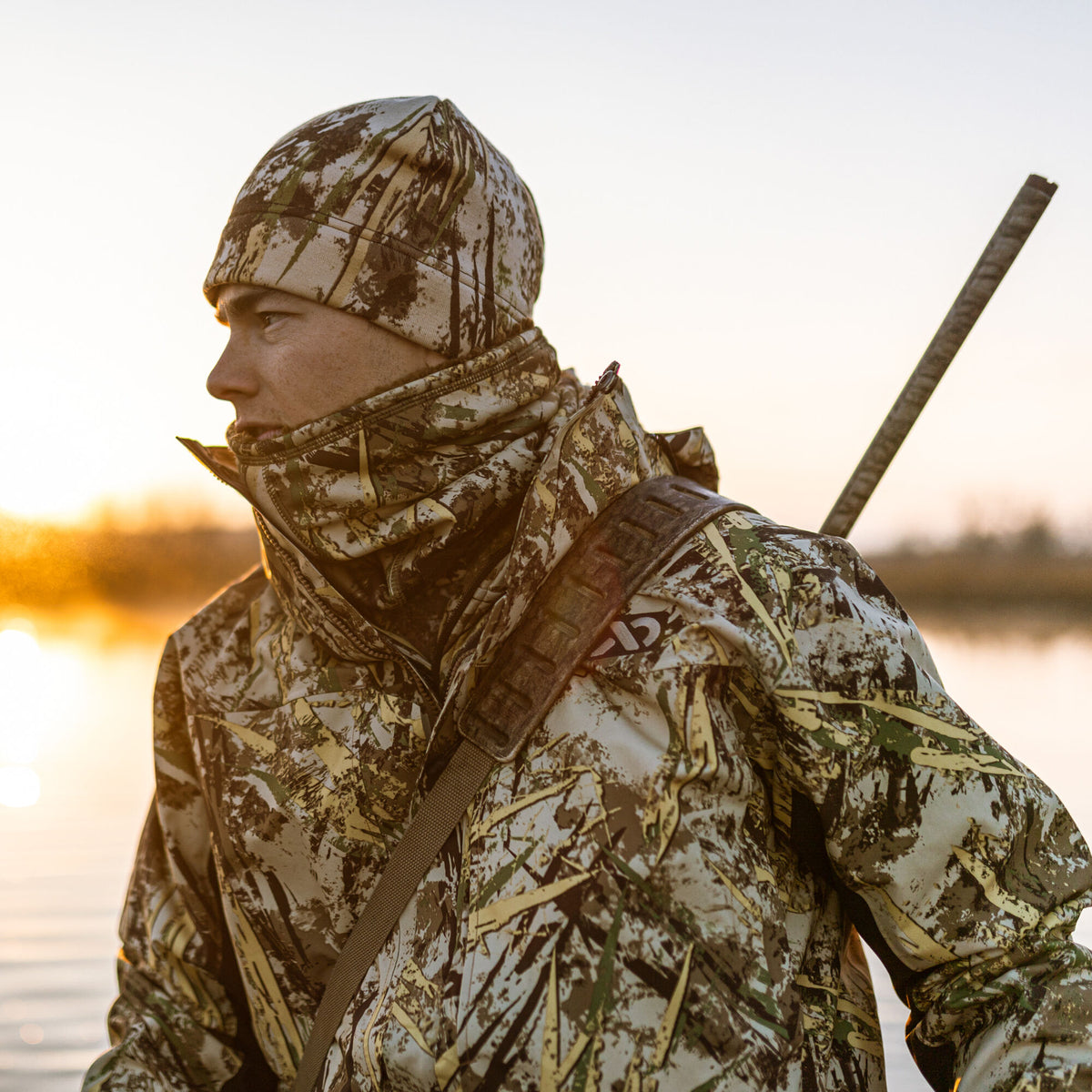 Man wearing a brown, green, and tan camouflage hunting jacket, beanie, and a rifle with a shoulder strap, facing left during sunset.