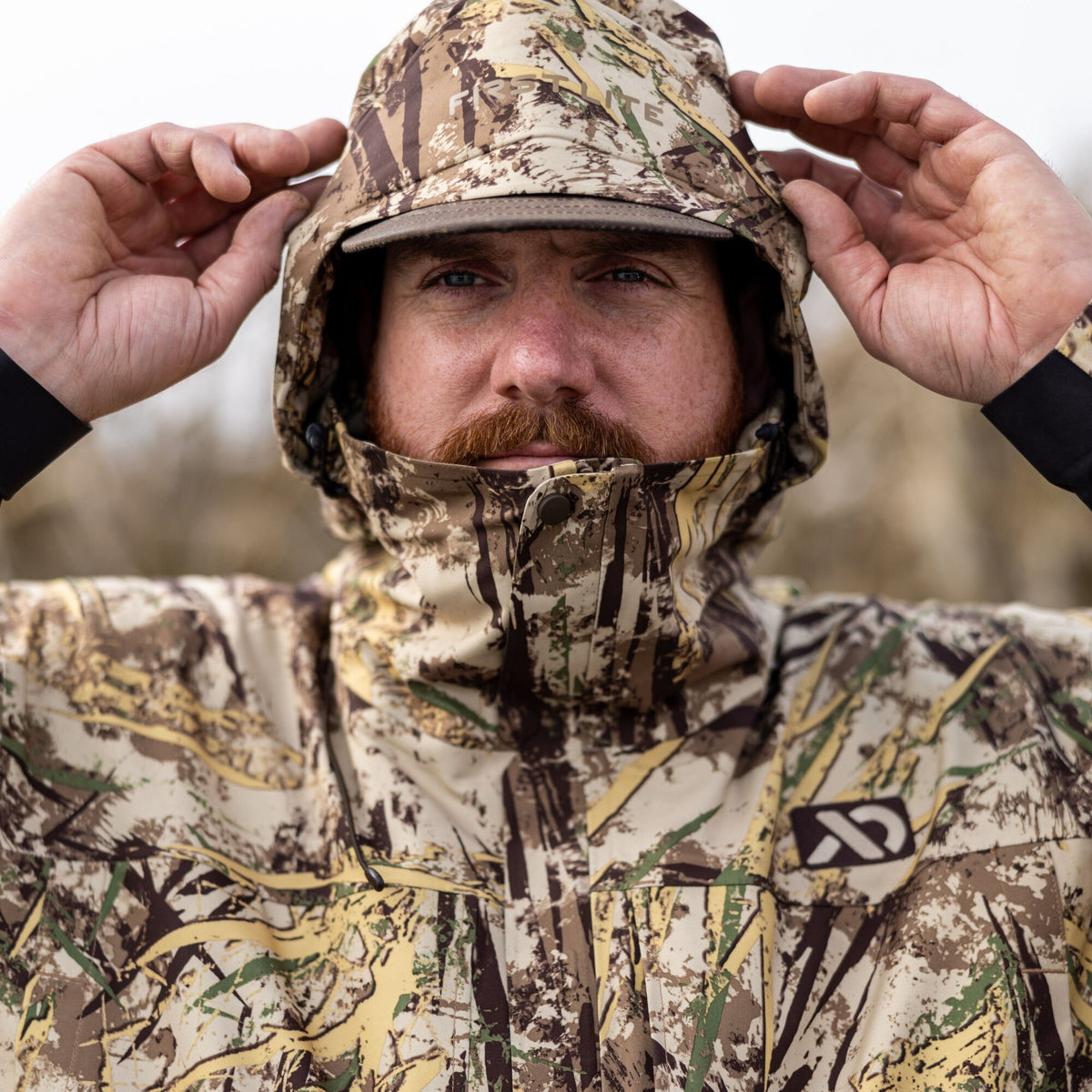 Man adjusting the hooded collar of a First Lite waterfowl camouflage jacket with a visible logo.