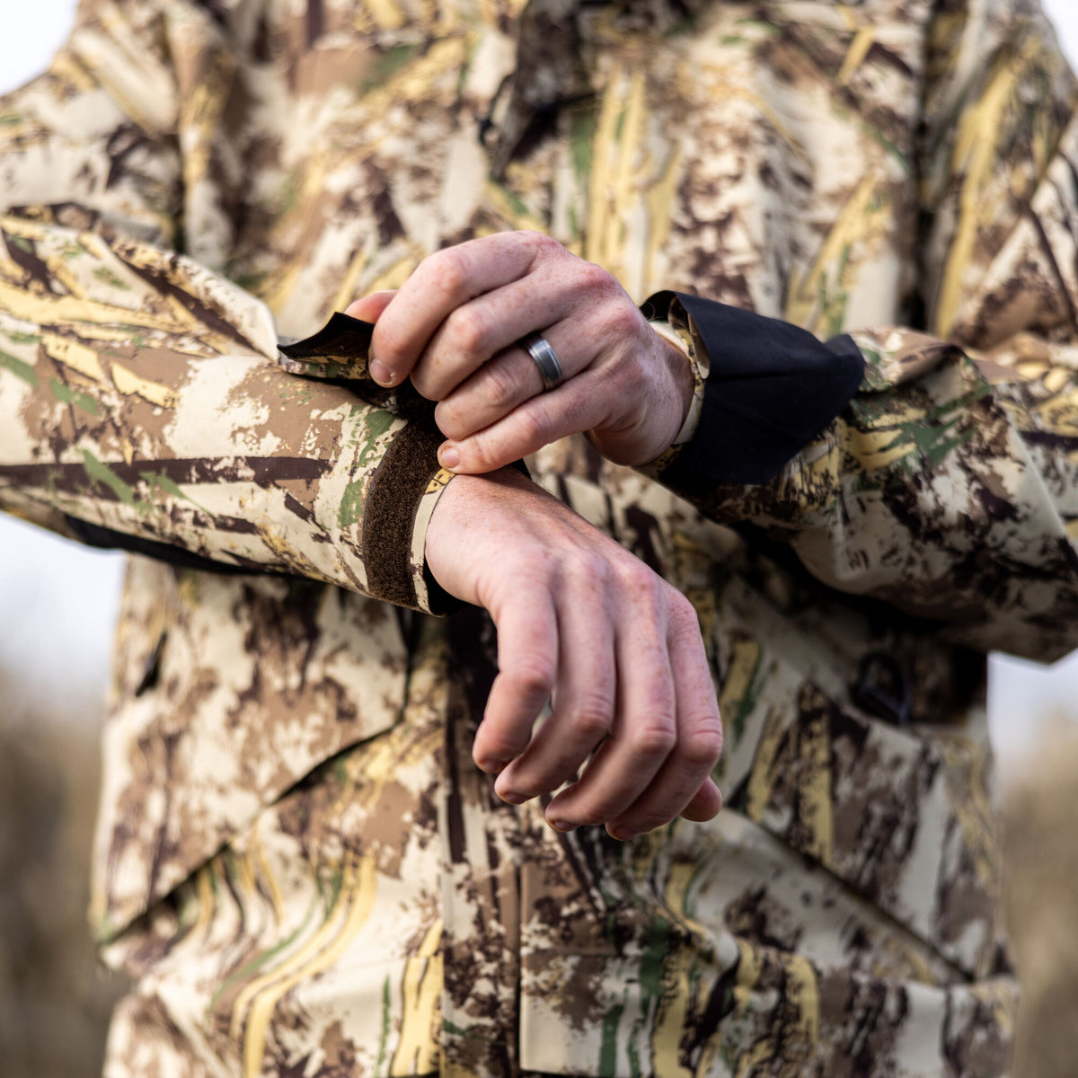 Close-up shows a person adjusting the hook-and-loop cuff of a tan, brown, and green camouflage jacket, revealing an inner black cuff.