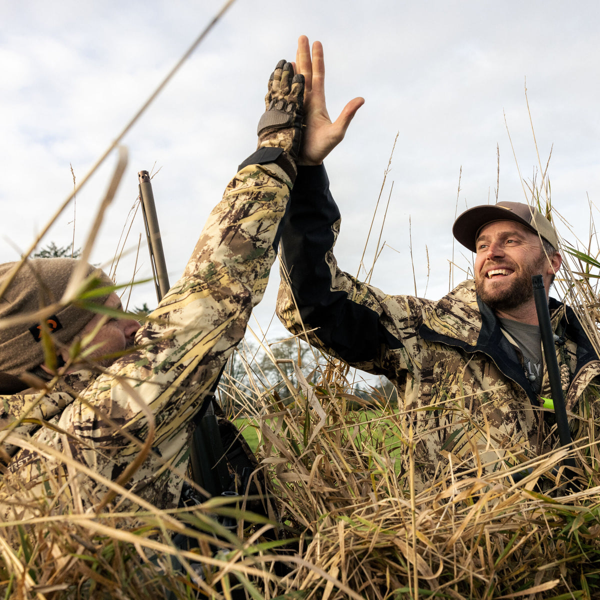 Two hunters in multi-color camouflage jackets with black side panels high-five in a tall grassy field, with a shotgun barrel visible.