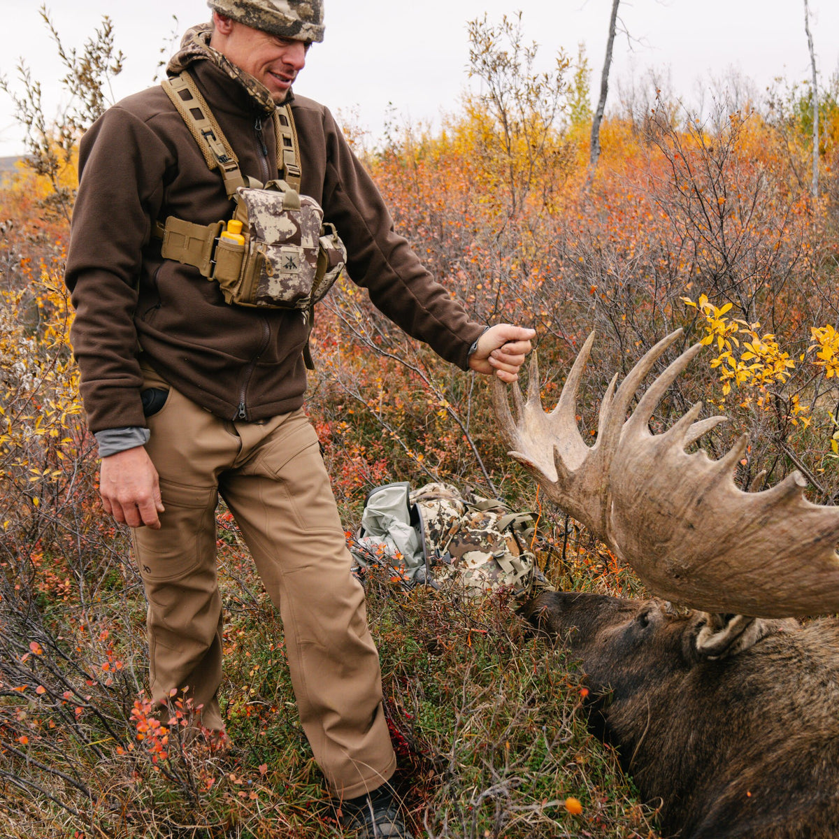 A smiling man in a brown fleece jacket, tan outdoor pants, and a camouflage chest rig holds the antlers of a large moose lying in an autumn forest.