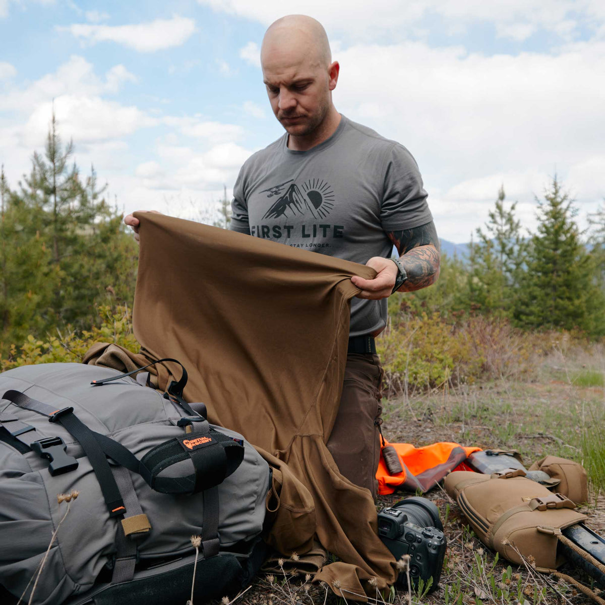 A man holds up a brown long-sleeve shirt, examining its fabric in an outdoor setting surrounded by hunting gear.