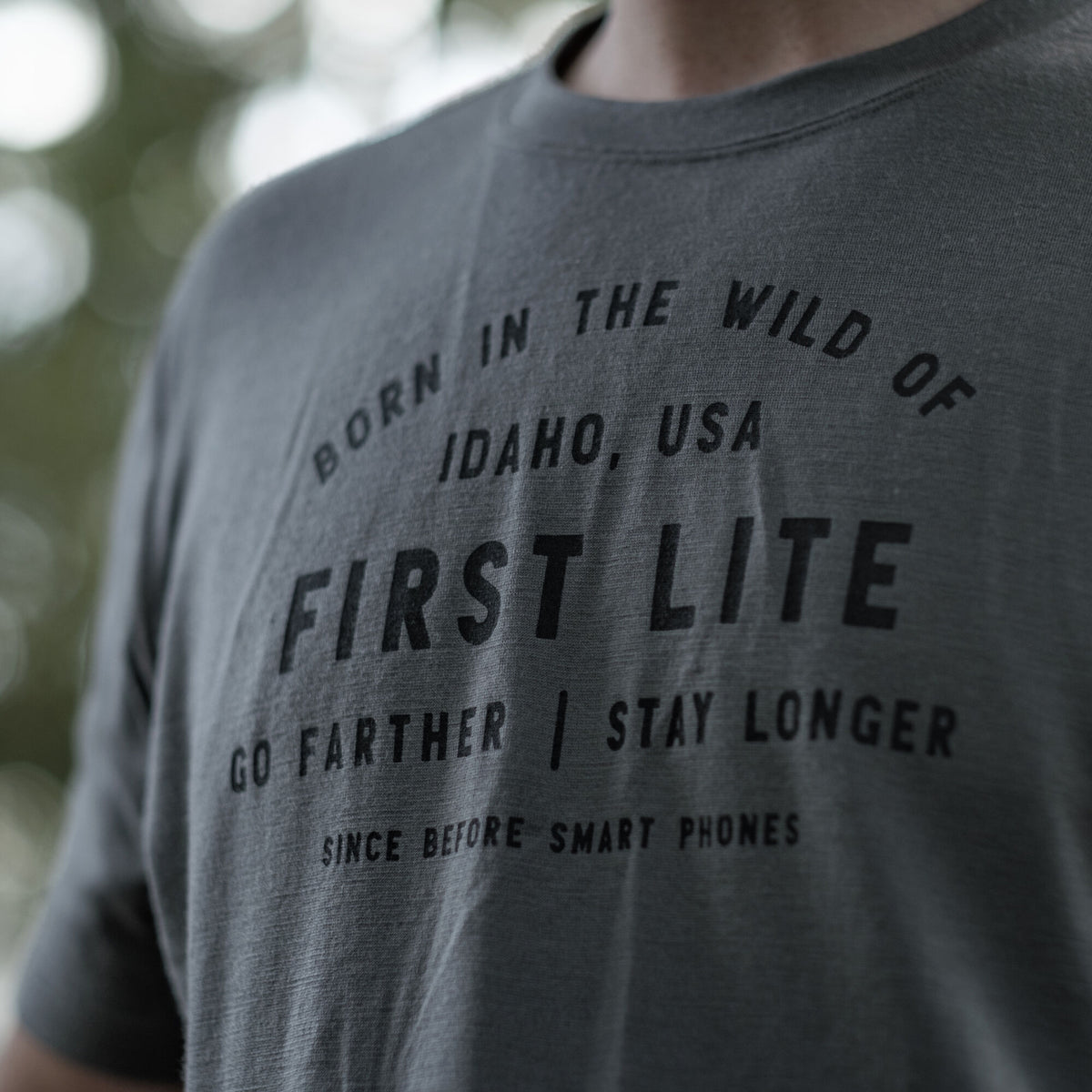 Dark grey t-shirt with "BORN IN THE WILD OF IDAHO, USA FIRST LITE GO FARTHER | STAY LONGER SINCE BEFORE SMART PHONES" printed in black text on the front.