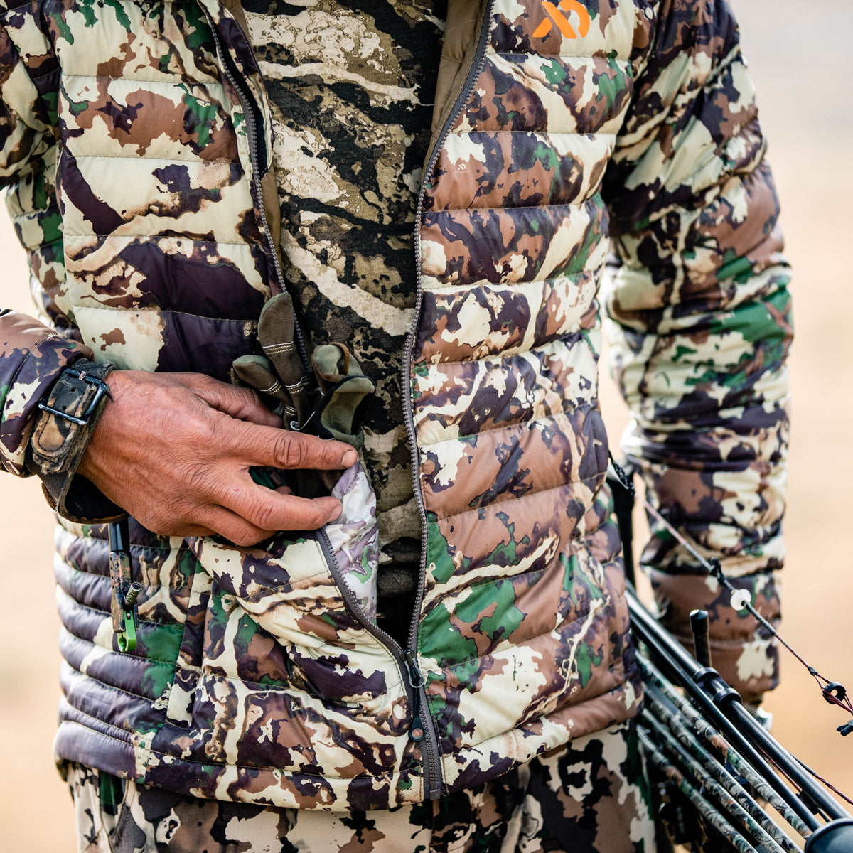 Brown, green, and tan camouflage quilted jacket with an orange logo, featuring a hand in a pocket and a hunting bow.