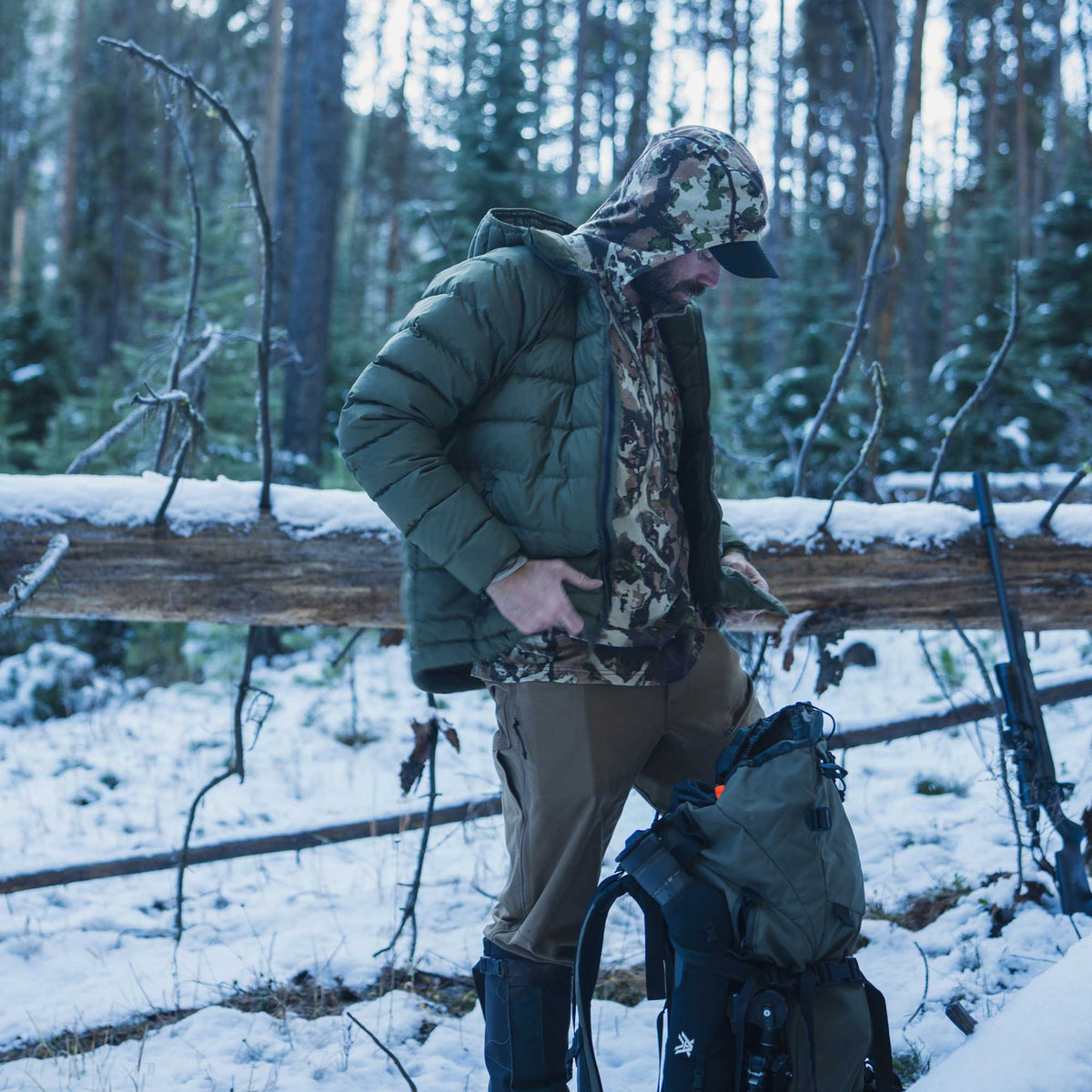 A man in a snowy forest wearing an olive green puffer jacket, camouflage hood, and brown pants, standing next to a large green and black backpack.