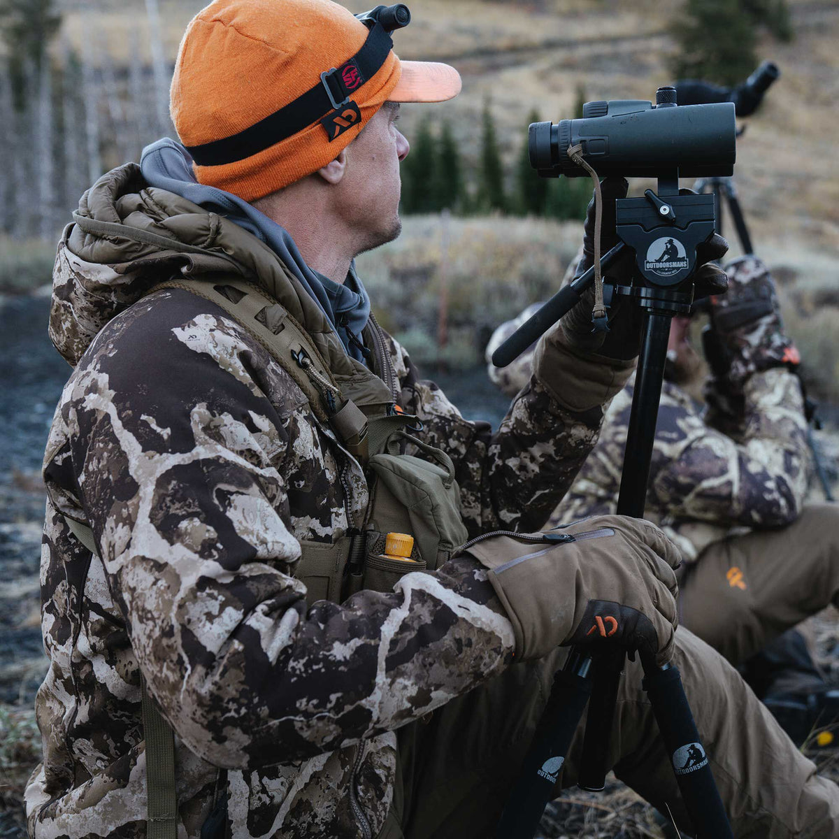 A person in an orange beanie with a headlamp and camouflage jacket holds a black spotting scope on a black Outdoorsmans tripod.