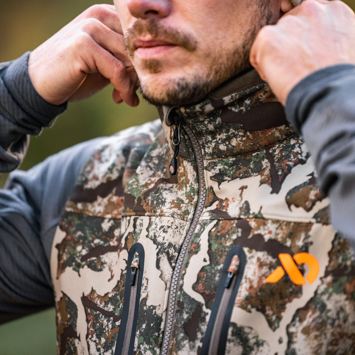 A man adjusts his camouflage vest, featuring a zippered front, two zippered pockets, and an orange logo.