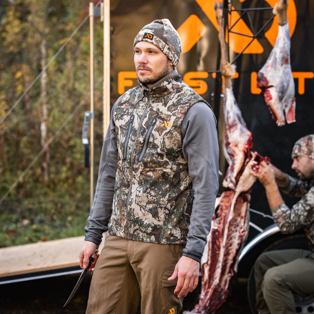 A man wears a camouflage hunting vest with zippered pockets, a gray long-sleeve shirt, brown pants, and a camouflage beanie with an orange logo.