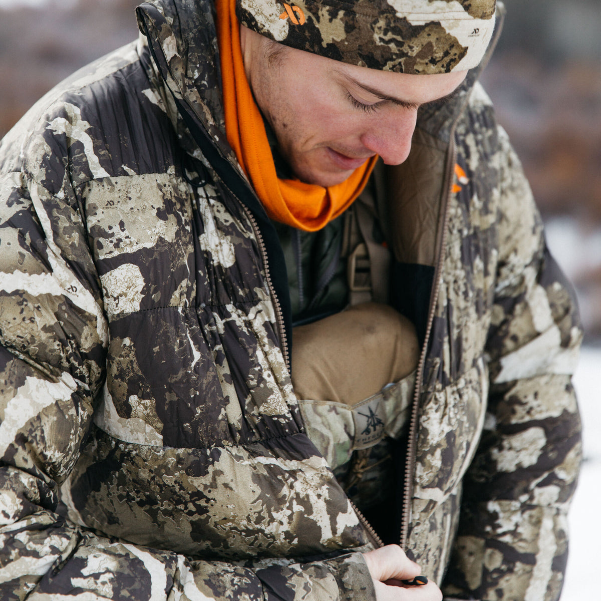 A person zips up a First Lite camouflage puffy jacket, revealing an orange neck gaiter and inner layers.
