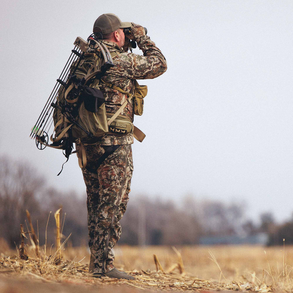 A hunter in multi-color camouflage clothing and a tan backpack with an attached compound bow looks through binoculars.