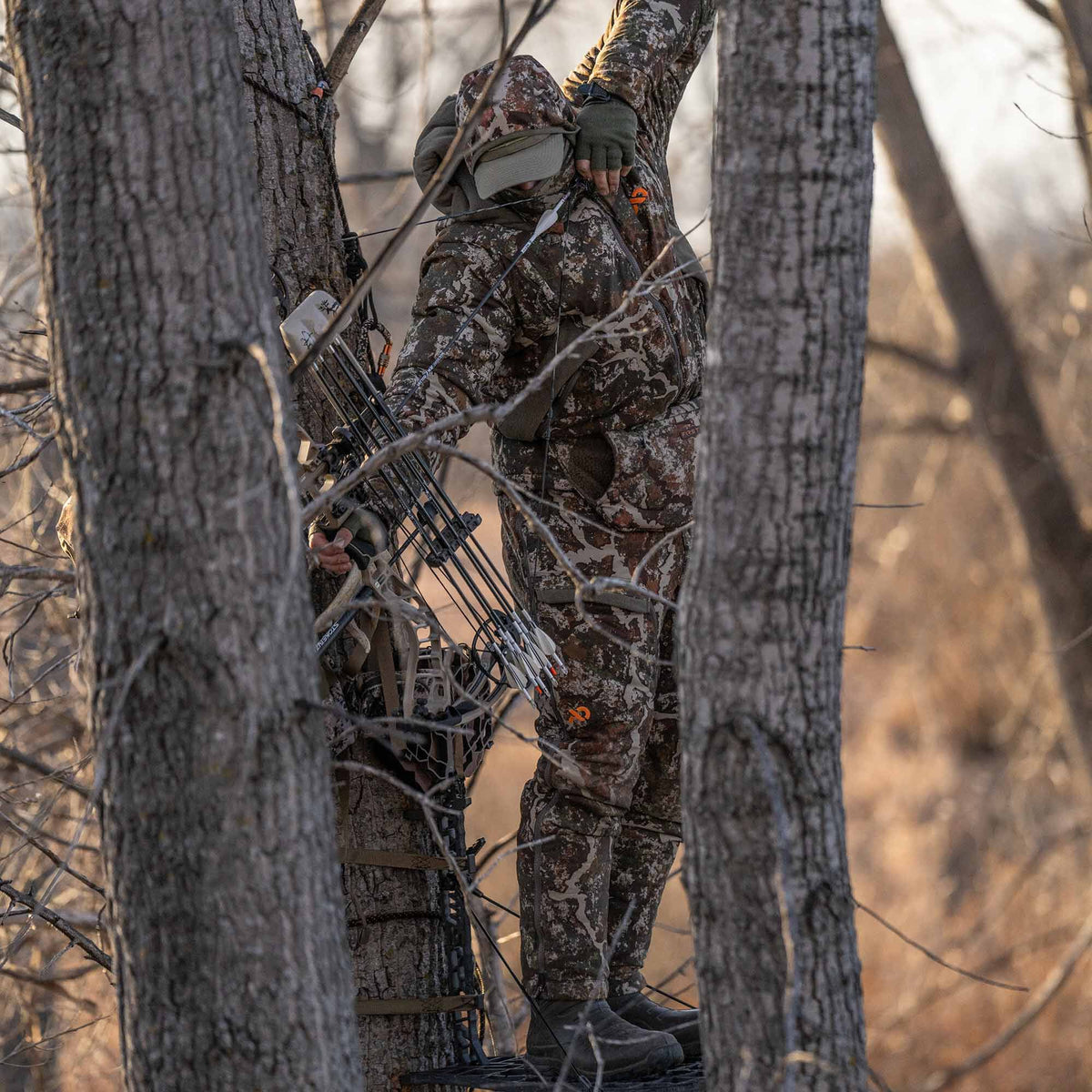 A hunter in full brown camouflage gear with an orange logo, holding a bow with quiver, standing in a tree stand.