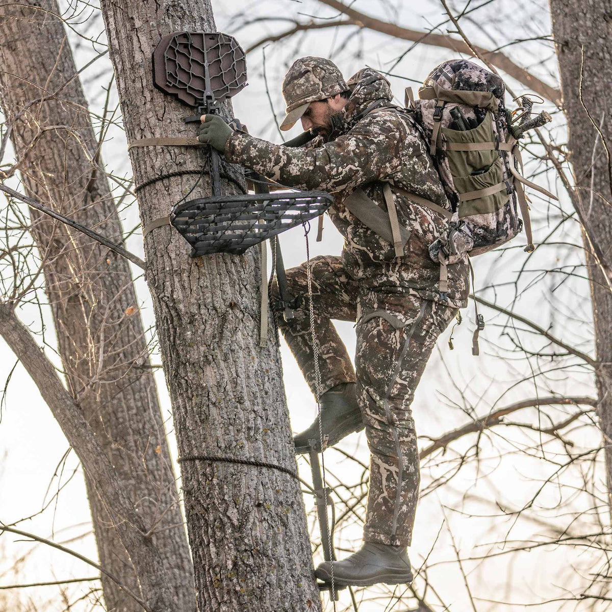 Dark grey tree stand with a web-patterned seat and climbing sticks, being used by a camouflage-clad hunter ascending a tree.
