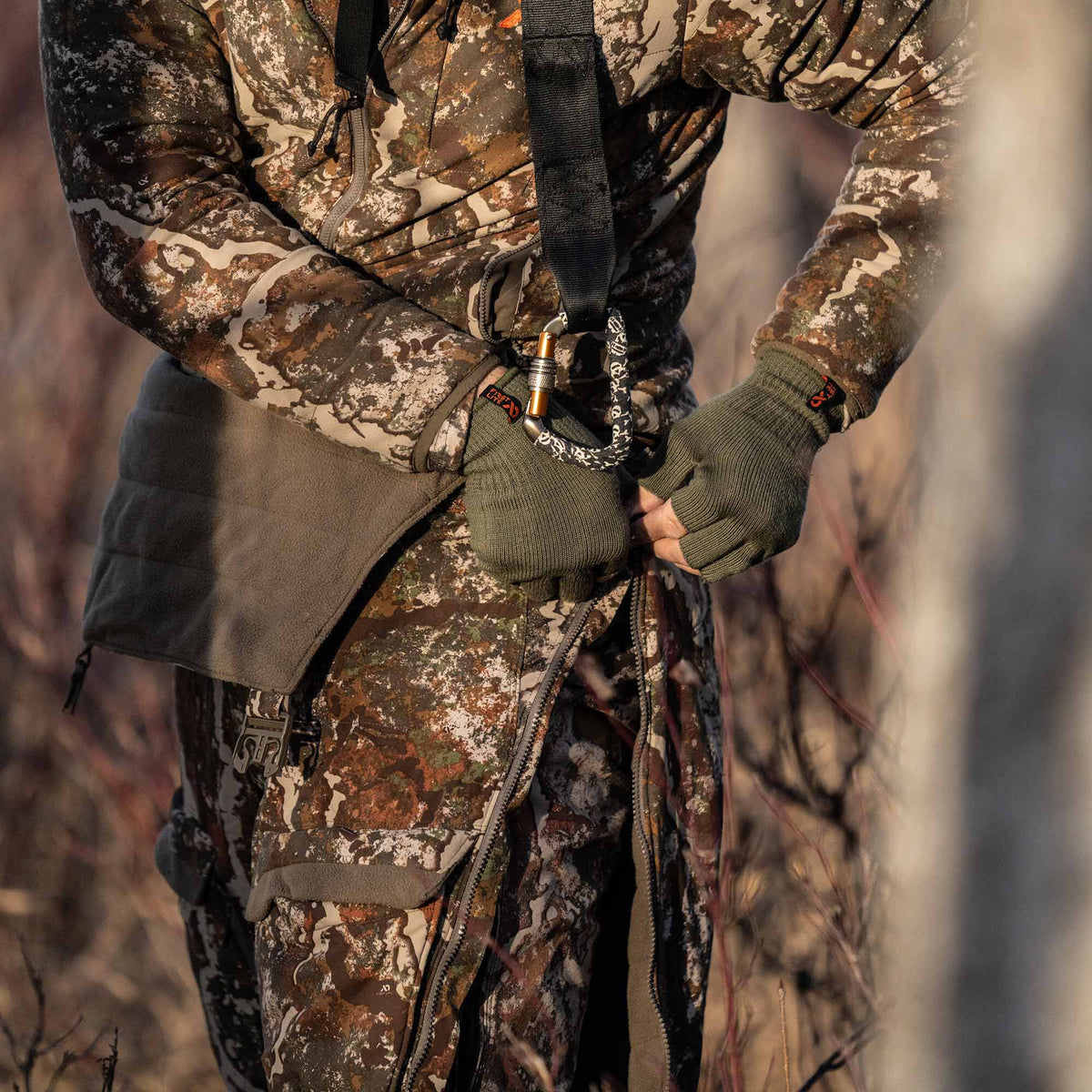 A person in brown, green, and tan camouflage hunting jacket and pants adjusts a pant side zipper with olive green fingerless gloves, with a hip pad and carabiner also visible.