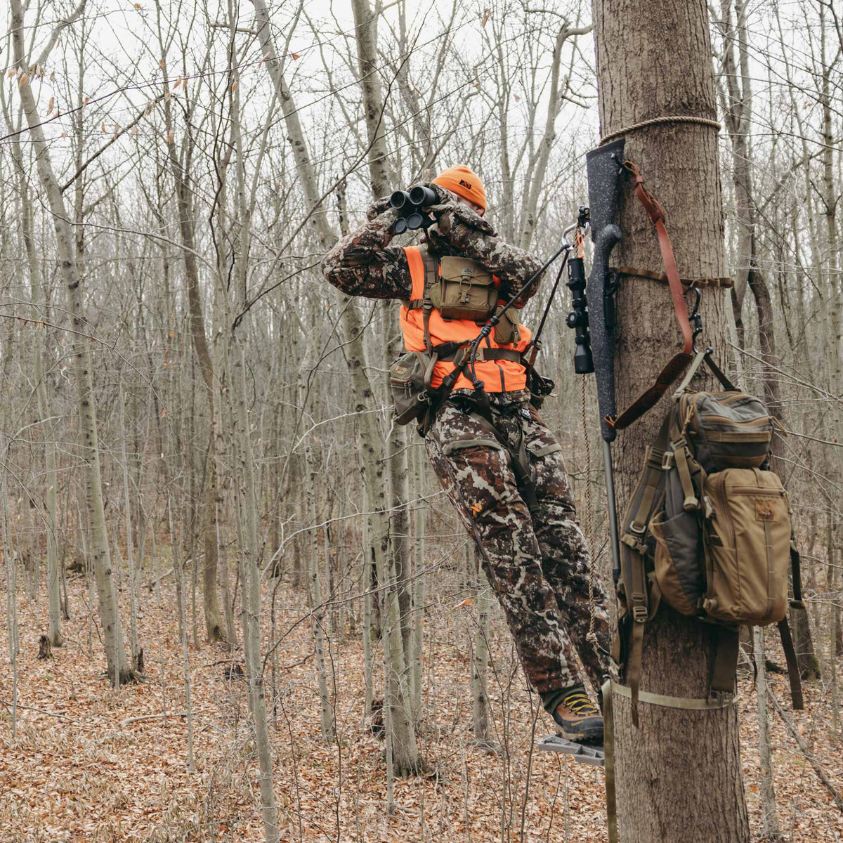 A hunter wearing a camouflage suit and orange safety vest sits elevated in a tree stand, scanning a leafless forest with binoculars while a rifle and tan backpack are secured to the tree.