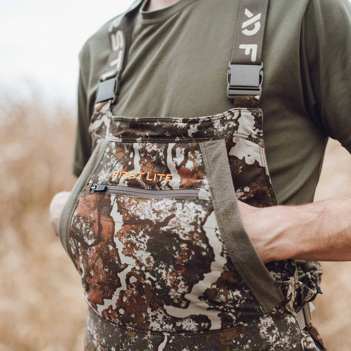Brown, green, and white camouflage hunting bibs featuring an orange First Lite logo, a zippered chest pocket, and side hand warmer pockets.