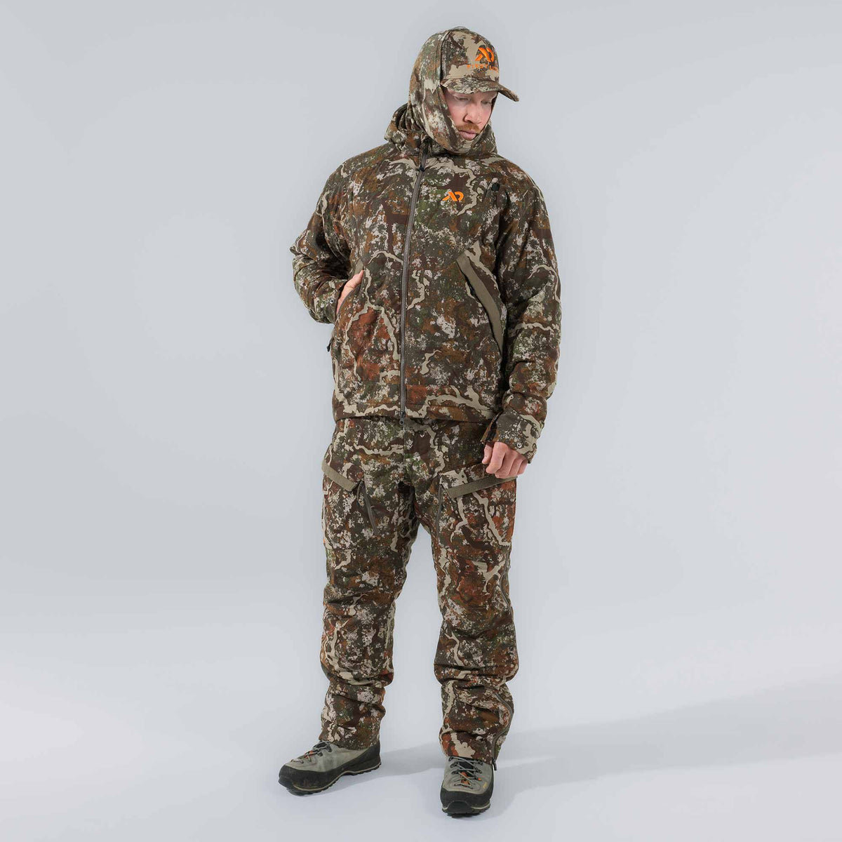 A man wearing a brown and green camouflage hooded hunting jacket with an orange logo, matching pants with suspenders, and a baseball cap.
