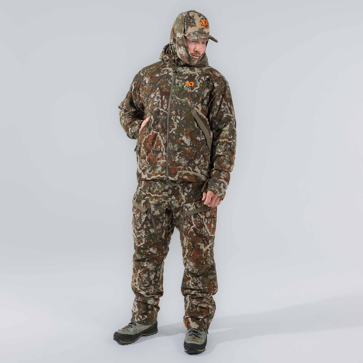 Man in a full camouflage hunting suit, featuring a hooded, full-zip jacket with an orange logo, pants with cargo pockets, and a matching cap.
