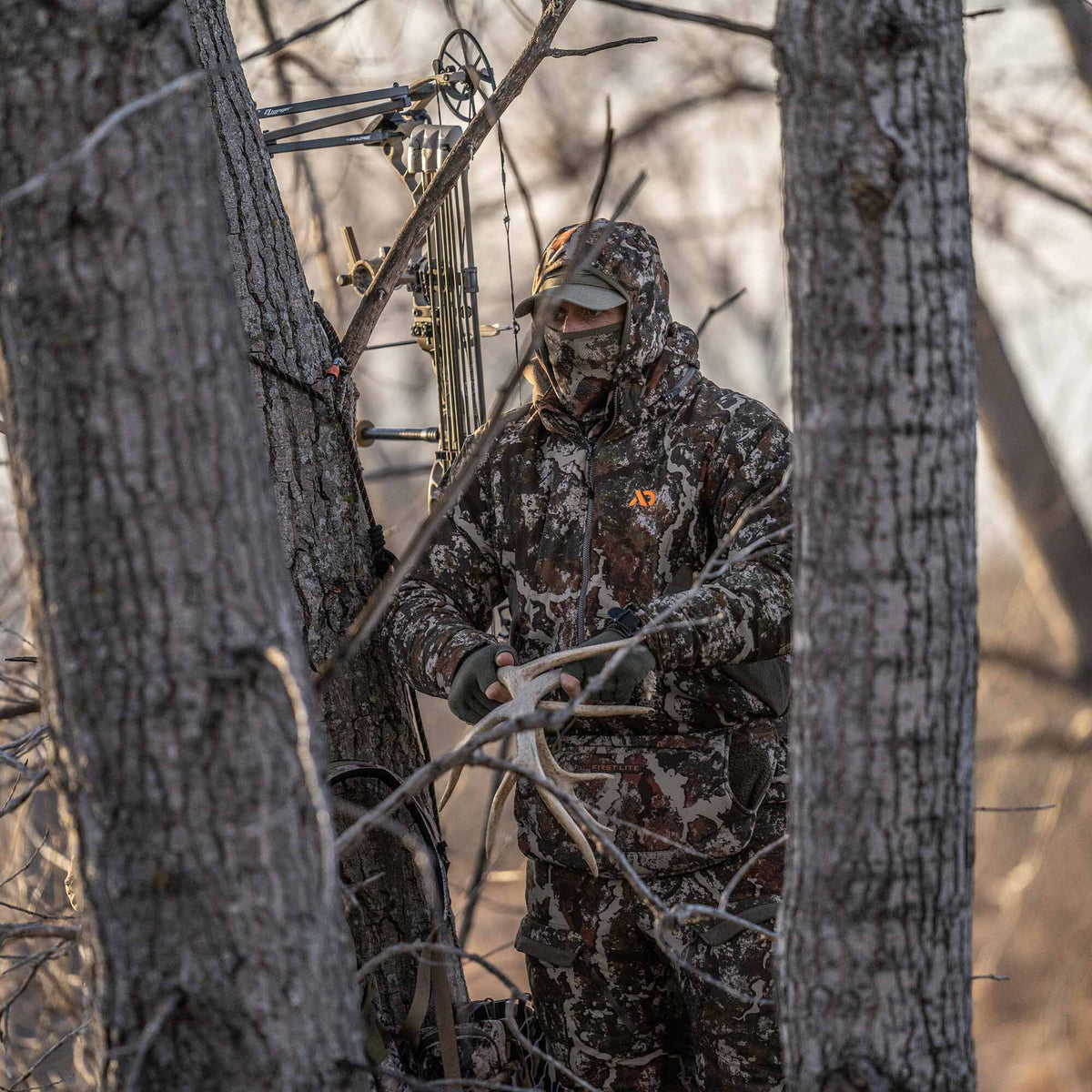 Hunter in First Lite brown and grey camouflage jacket, balaclava, and gloves holds a deer antler by a bow in a forest.