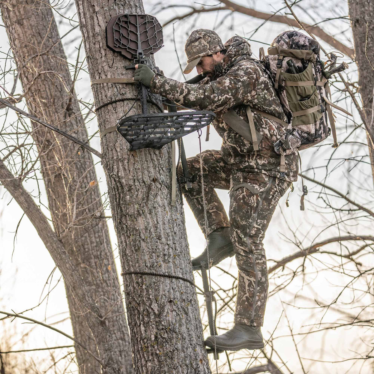 Hunter in camouflage uses a dark brown climbing tree stand with a webbed seat and platform to ascend a tree.