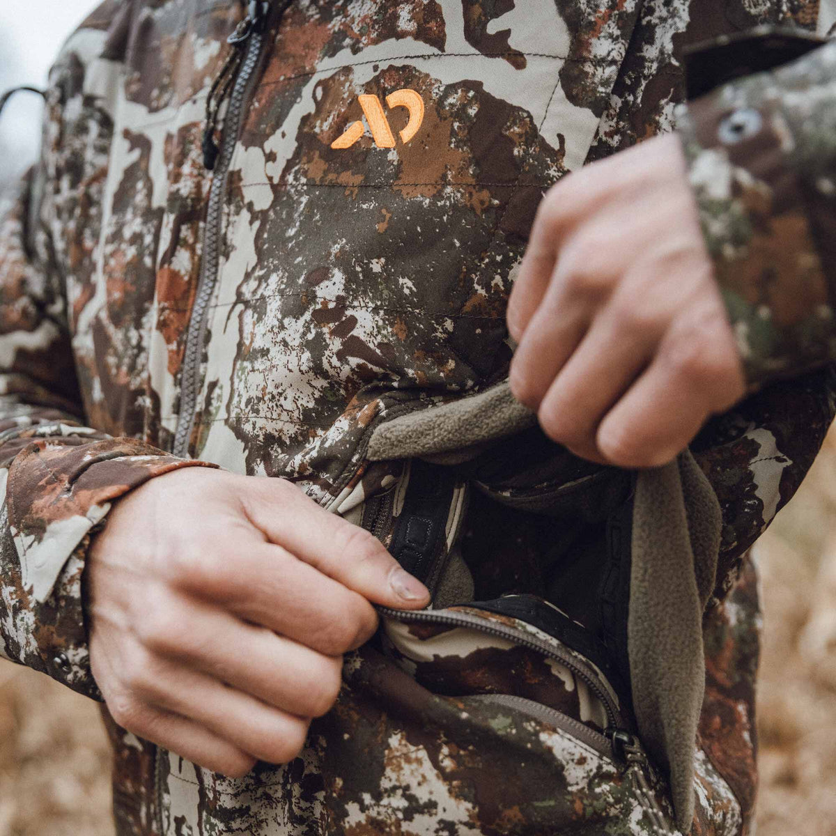 Hands open a zippered, fleece-lined pocket on a brown, green, and white camouflage jacket featuring an orange chest logo.