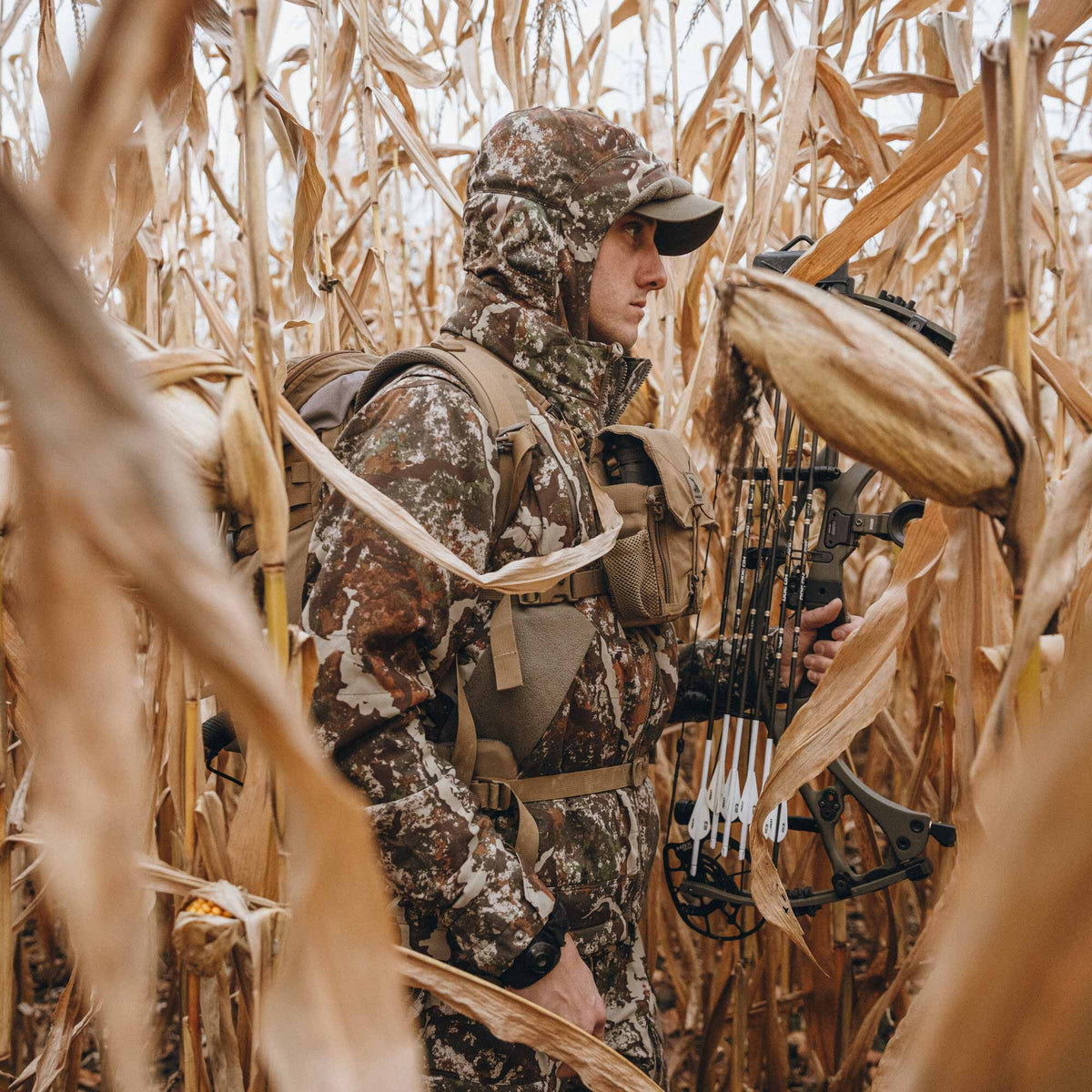 A person wears a multi-colored camouflage hooded hunting suit with a backpack, chest pouch, and compound bow with arrows in a dry cornfield.