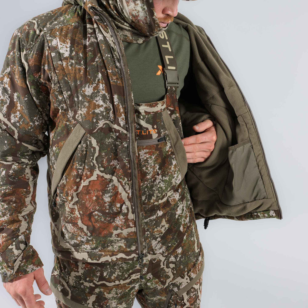 Person wearing an open brown and green camouflage hunting jacket and matching bibs, revealing olive green fleece lining, an olive green t-shirt, and suspenders.