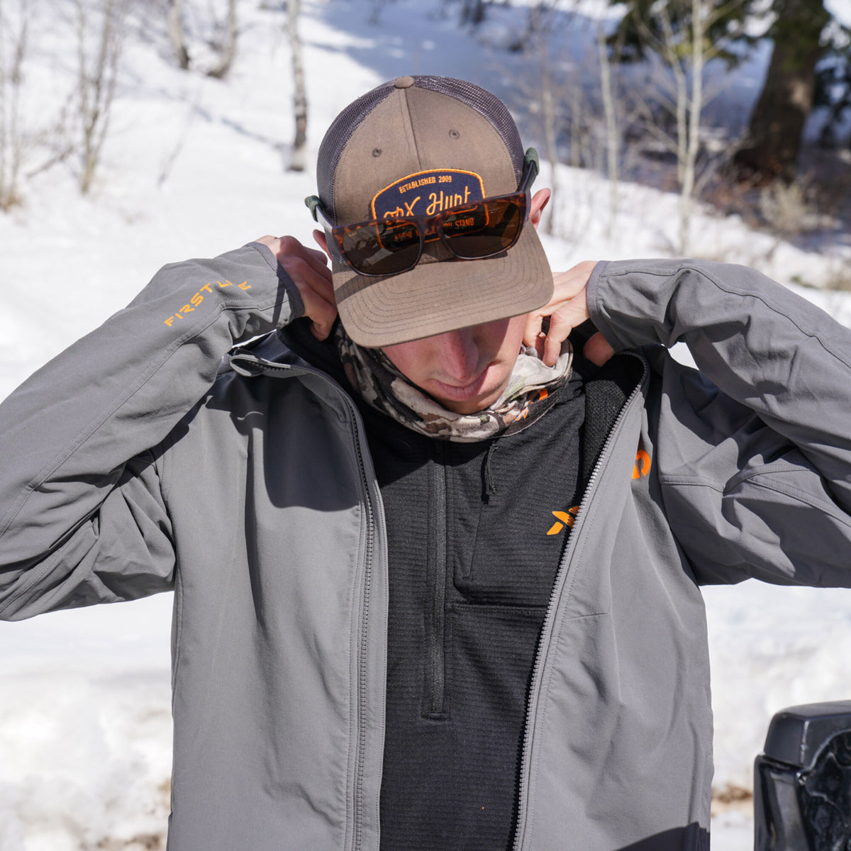 A person wears a gray full-zip jacket with orange "FIRST LITE" text, over a dark gray quarter-zip pullover with an orange logo and a camouflage neck gaiter, topped by a brown trucker hat with an "OX Hunt" patch and tortoiseshell sunglasses, in a snowy landscape.