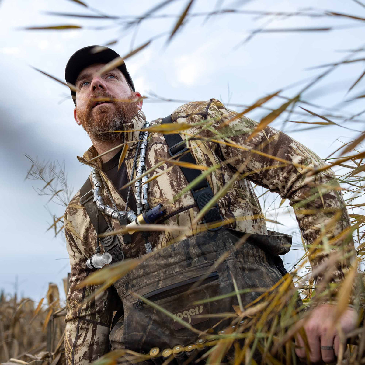 Man in a brown and green camouflage hunting wader outfit, featuring a duck band lanyard, duck calls, and a Rogers logo.