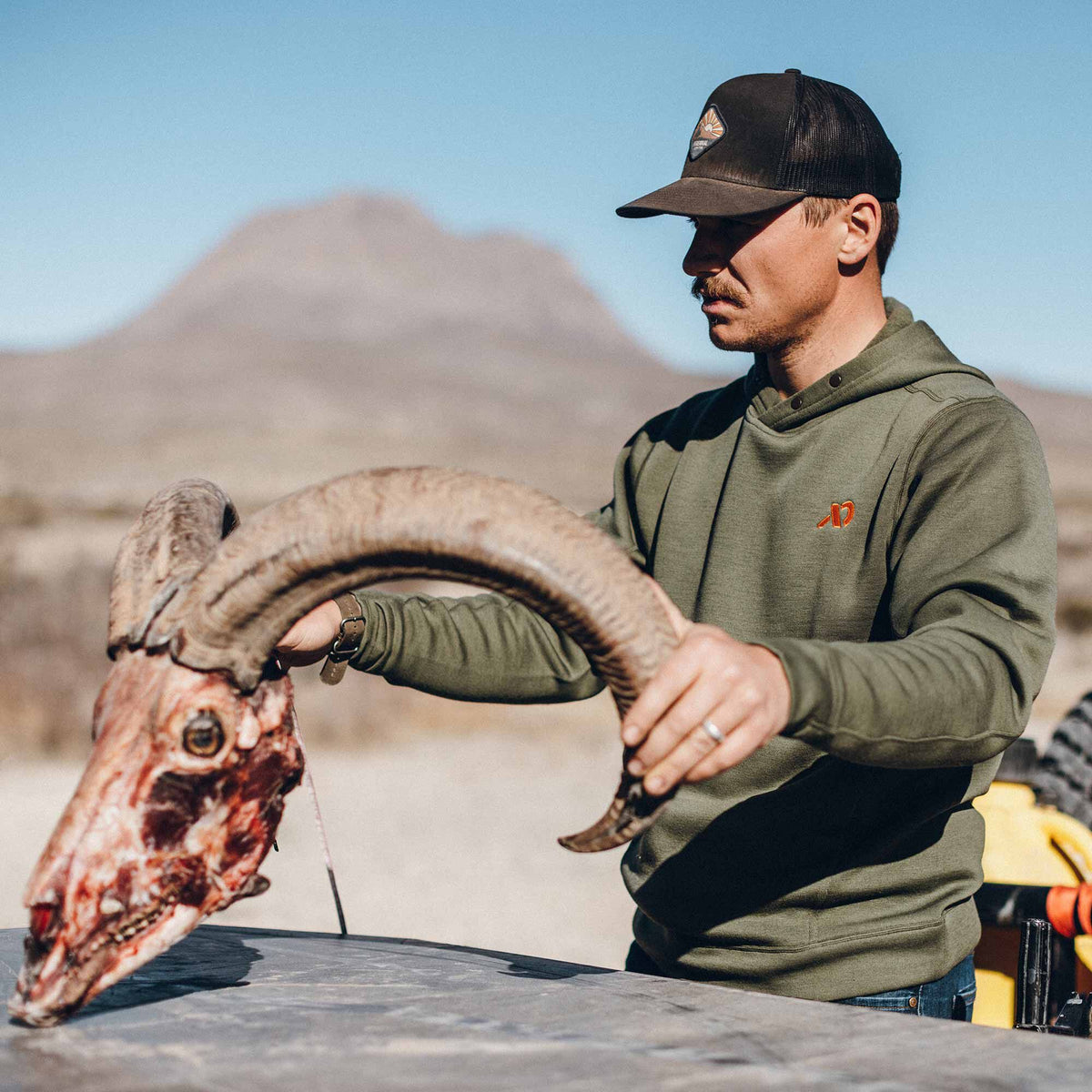 Man wearing an olive green pullover hoodie with snap placket and orange chest logo, holding a bighorn sheep skull.