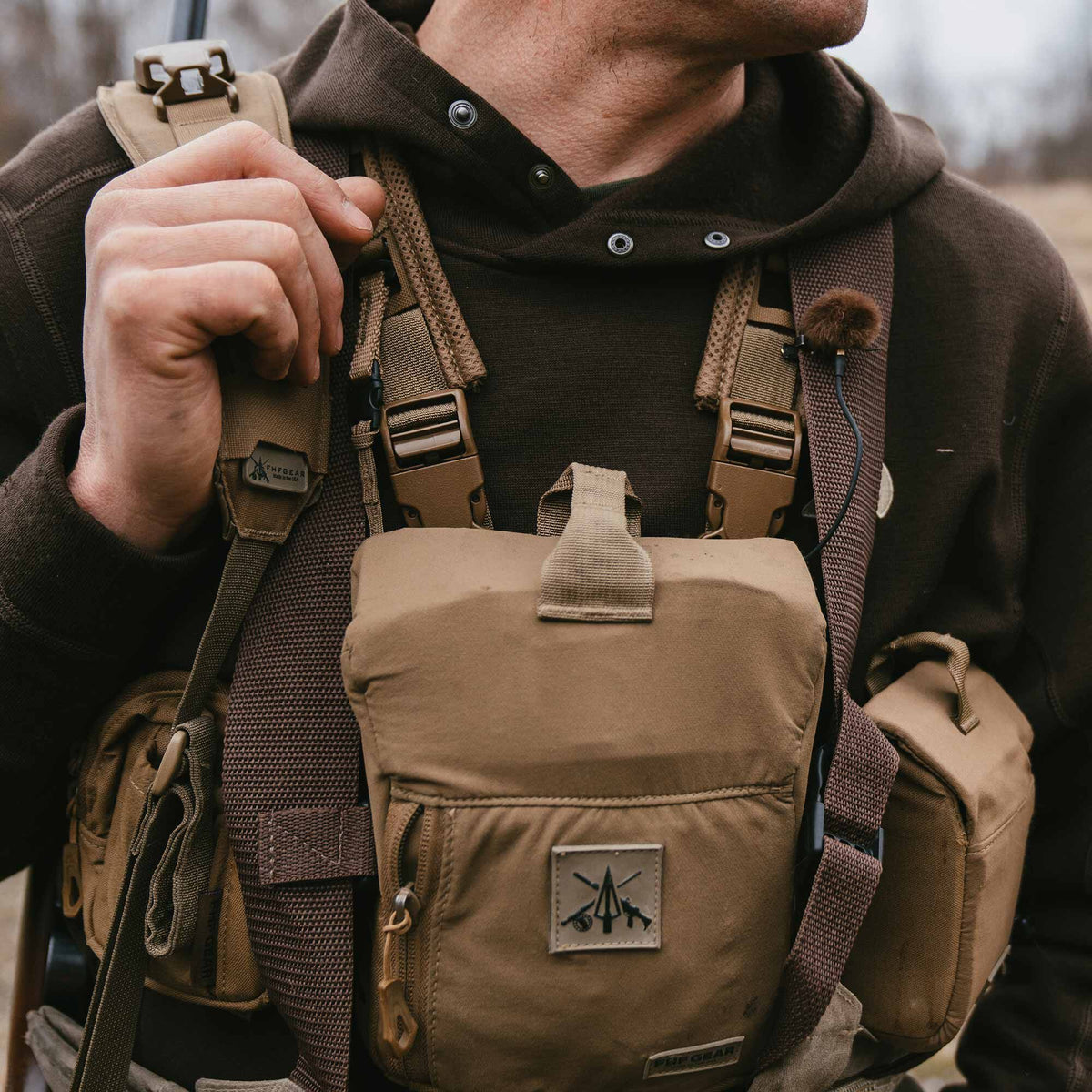 Tan binocular harness featuring a central logo patch, side pouches, and dark brown mesh-backed straps, worn by a person.